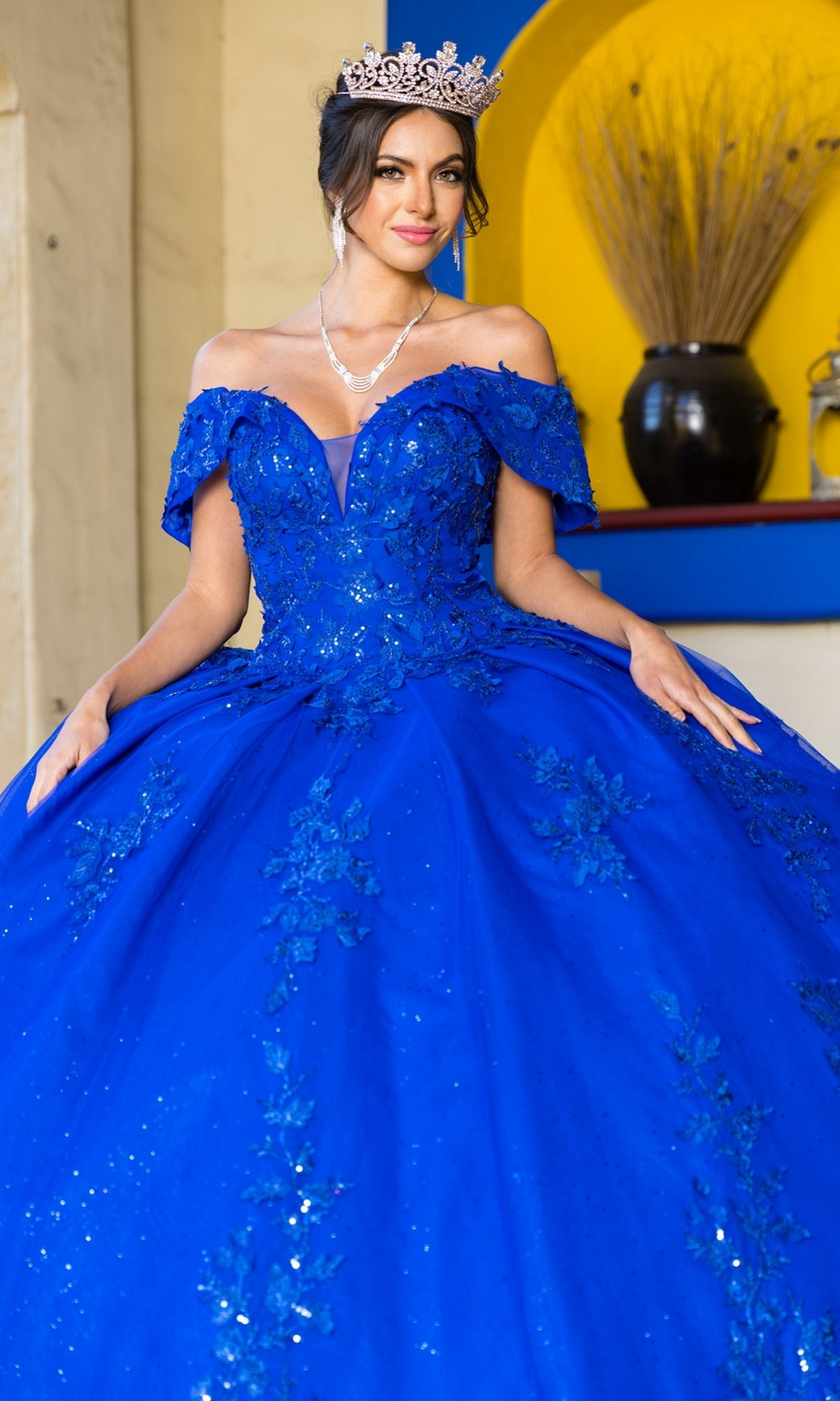 A woman in the Quinceanera Ball Gown 8045J with glitter lace embroidery and a tiara stands indoors before a yellow and blue alcove.